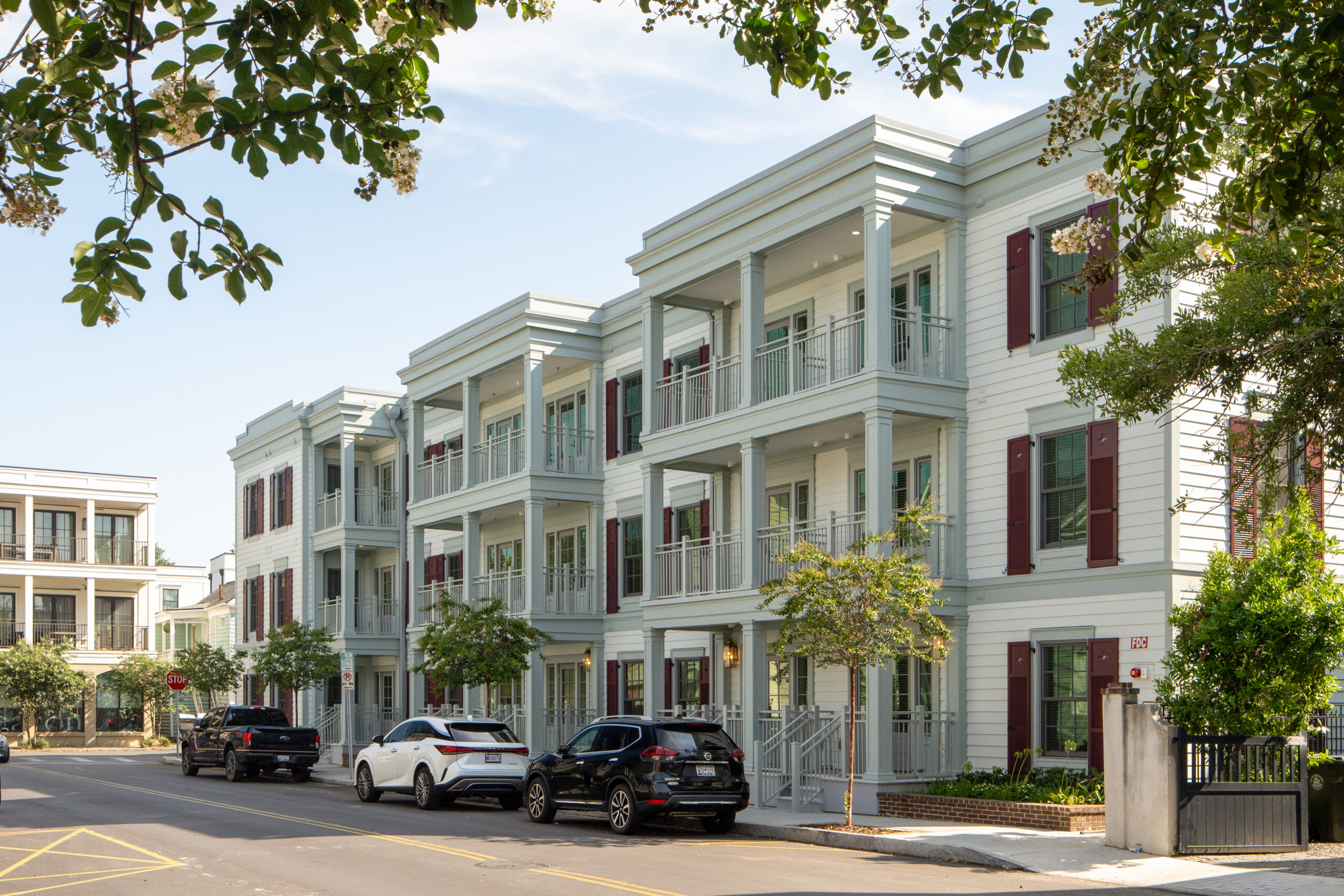 Exterior view of a modern three-story apartment community with white siding, balconies, burgundy shutters, and tree-lined streets.