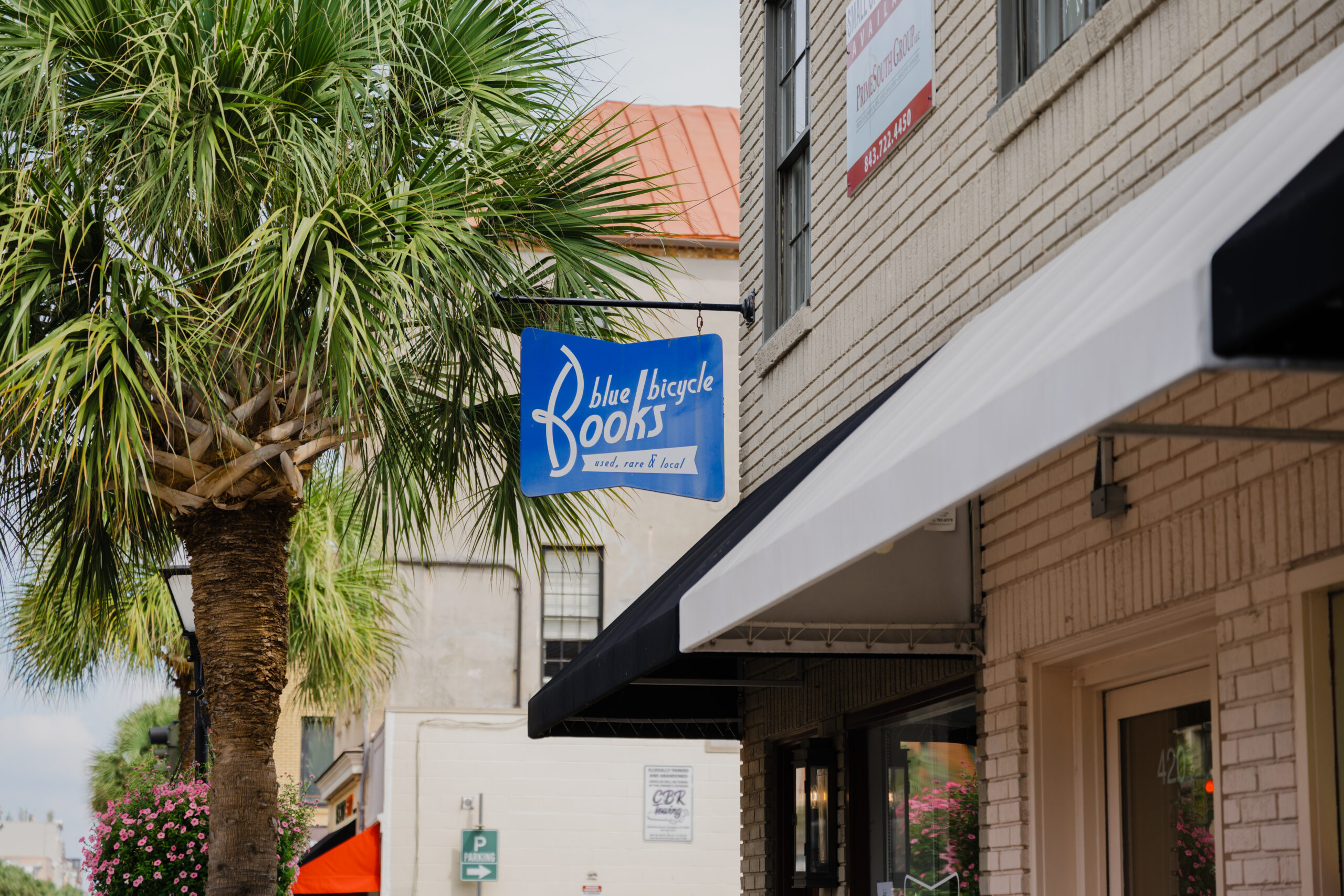A sun-drenched outdoor view of a city sidewalk featuring a prominent blue hanging sign for 