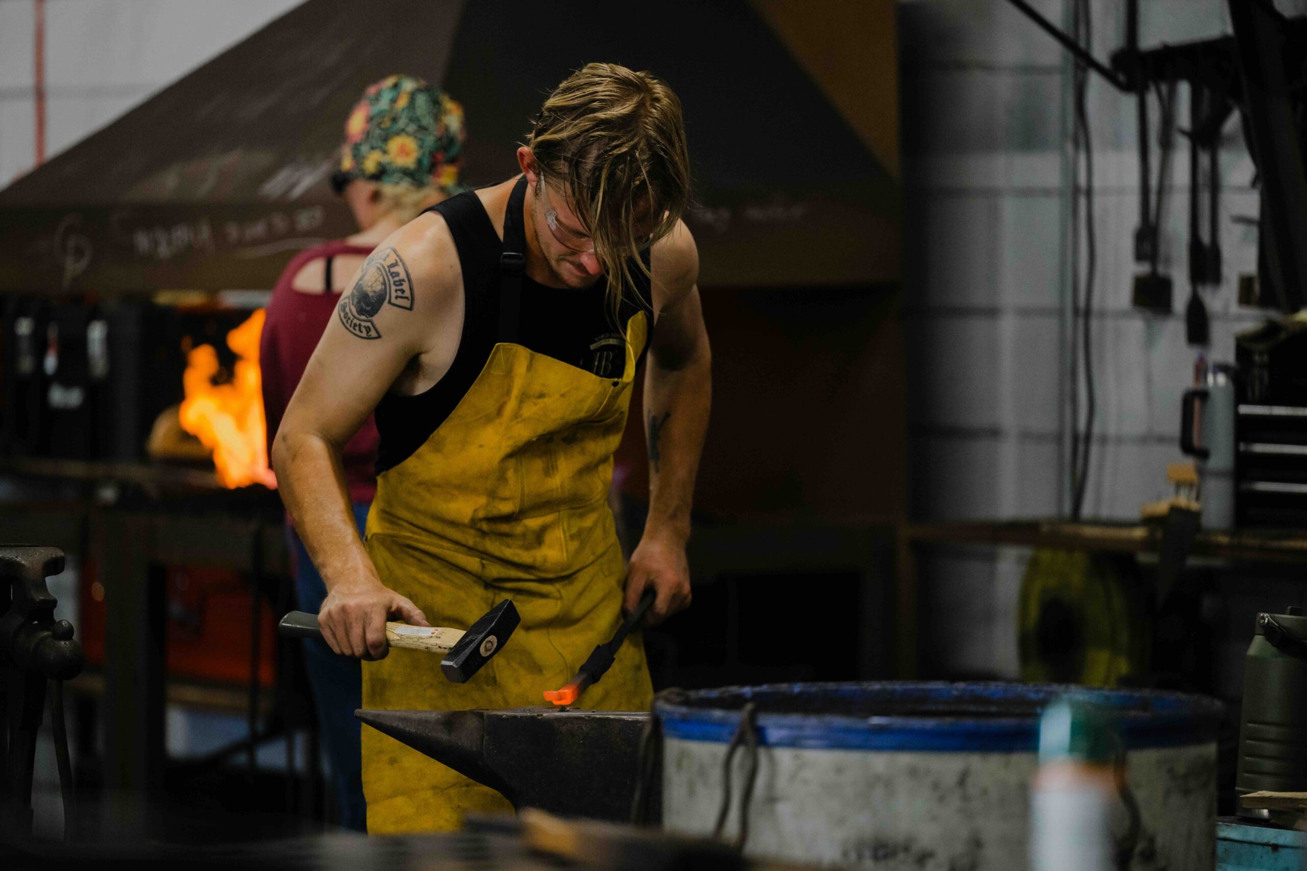 A high-action shot of a male blacksmith at work in a dark, industrial forge. The man, with blonde hair and a tattoo on his right shoulder, wears a heavy yellow protective apron over a black tank top and safety glasses. He is leaning over a large black anvil, holding a glowing orange-hot metal piece with tongs while preparing to strike it with a heavy blacksmithing hammer. In the soft-focus background, another worker is visible near a roaring orange fire from a furnace, and various metal-working tools hang on a white cinderblock wall. The lighting is dramatic, highlighting the sparks, the heat of the metal, and the intense physical effort of the craft.