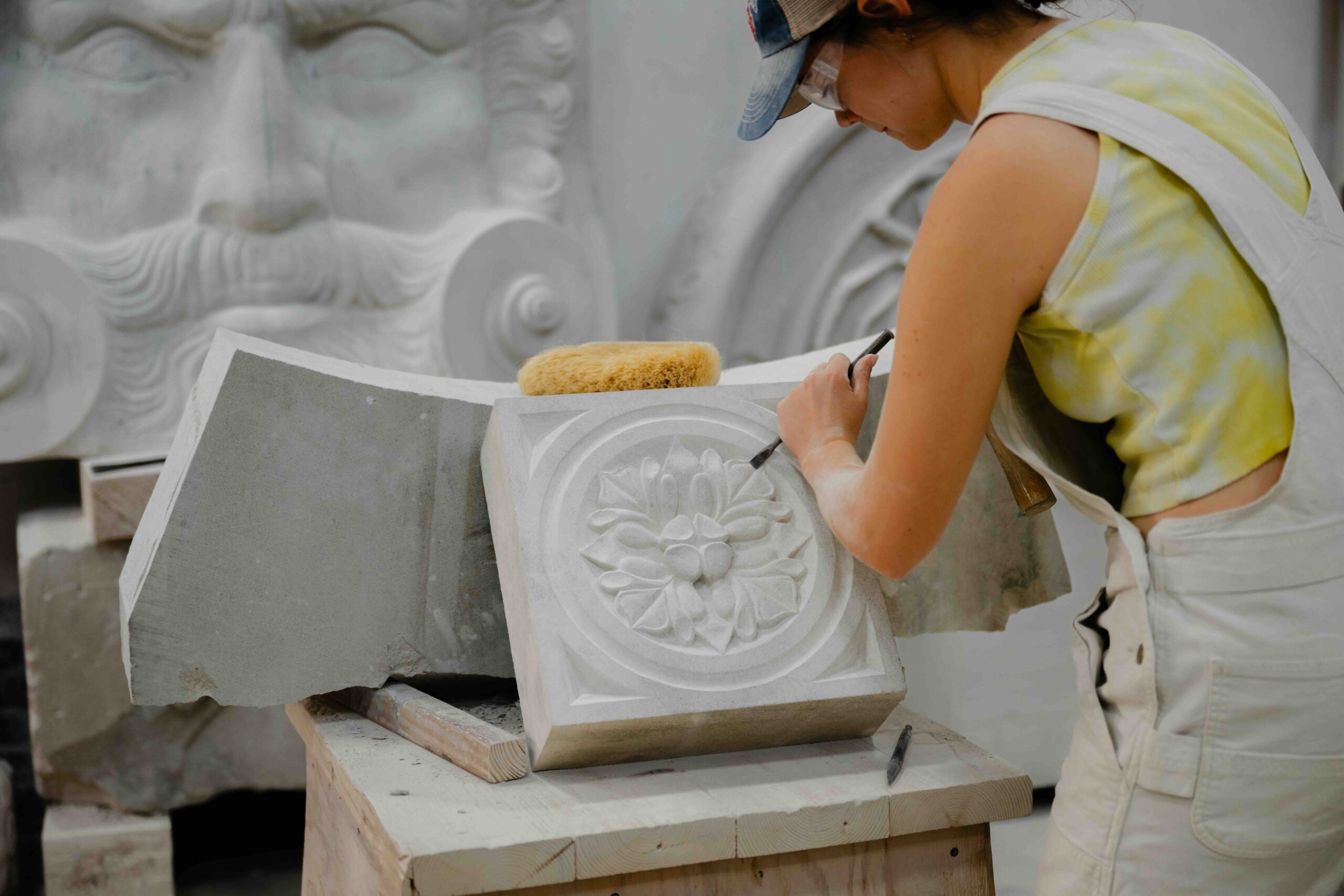 A close-up side profile of a female artisan working in a stone carving studio. She is wearing a yellow tie-dye tank top, cream-colored denim overalls, a baseball cap, and clear safety glasses. She is leaning over a wooden workbench, using a fine metal chisel and a wooden mallet to carve an intricate, symmetrical floral rosette into a square block of light grey stone. On the bench beside her work rests a natural sea sponge and another larger, curved piece of unfinished stone. In the soft-focus background, a massive, ornate classical stone face with a large mustache and swirling architectural details is visible, highlighting the scale and craftsmanship of the workshop environment.