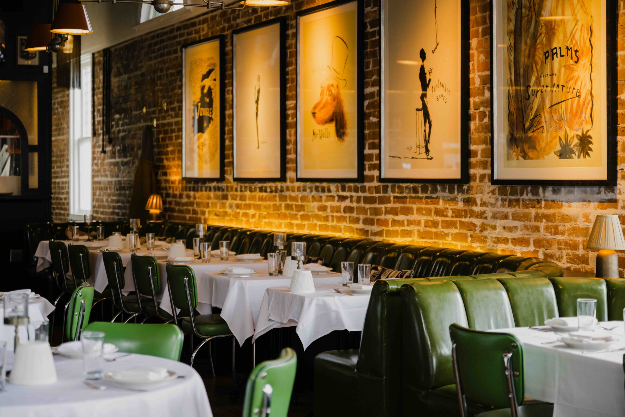 An interior wide shot of a dimly lit, upscale restaurant featuring a long row of dark green tufted leather banquette seating against an exposed red and tan brick wall. Each dining table is draped in crisp white linens and set with sparkling glassware, white napkins, and small shaded table lamps that cast a warm, golden glow across the room. Above the seating, a series of five large, vertically oriented framed art prints with whimsical yellow-toned illustrations are mounted in black frames. The foreground features additional round tables with green leather chairs, creating a sense of depth and a sophisticated, club-like atmosphere perfect for professional gatherings or celebratory dinners.