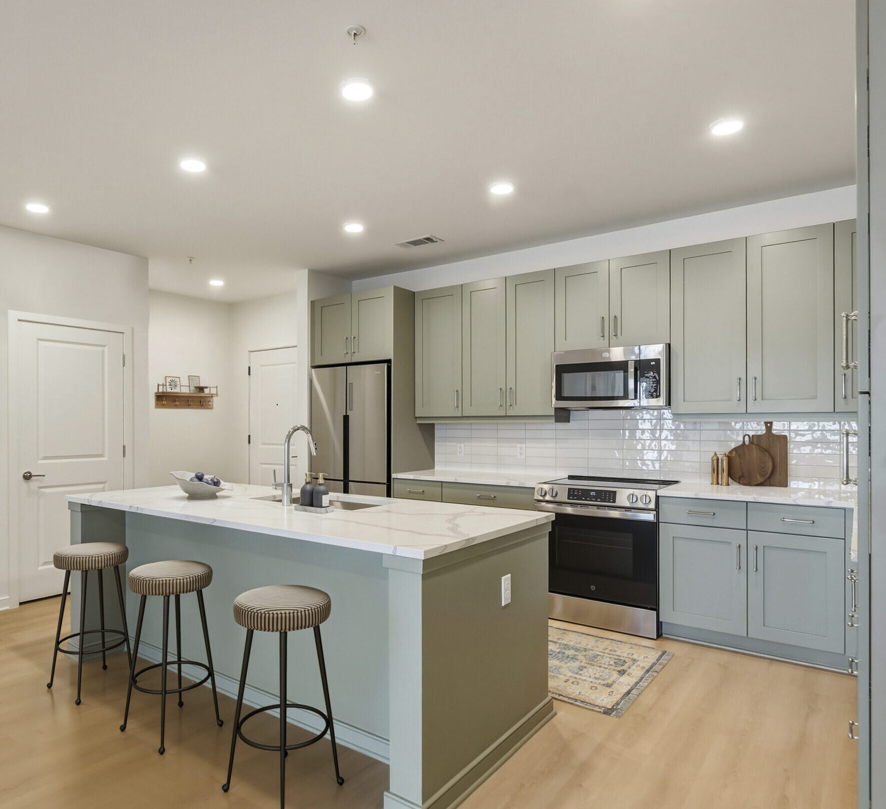Modern kitchen with sage green shaker cabinets, white quartz countertops, stainless steel appliances, subway tile backsplash, and light wood flooring.