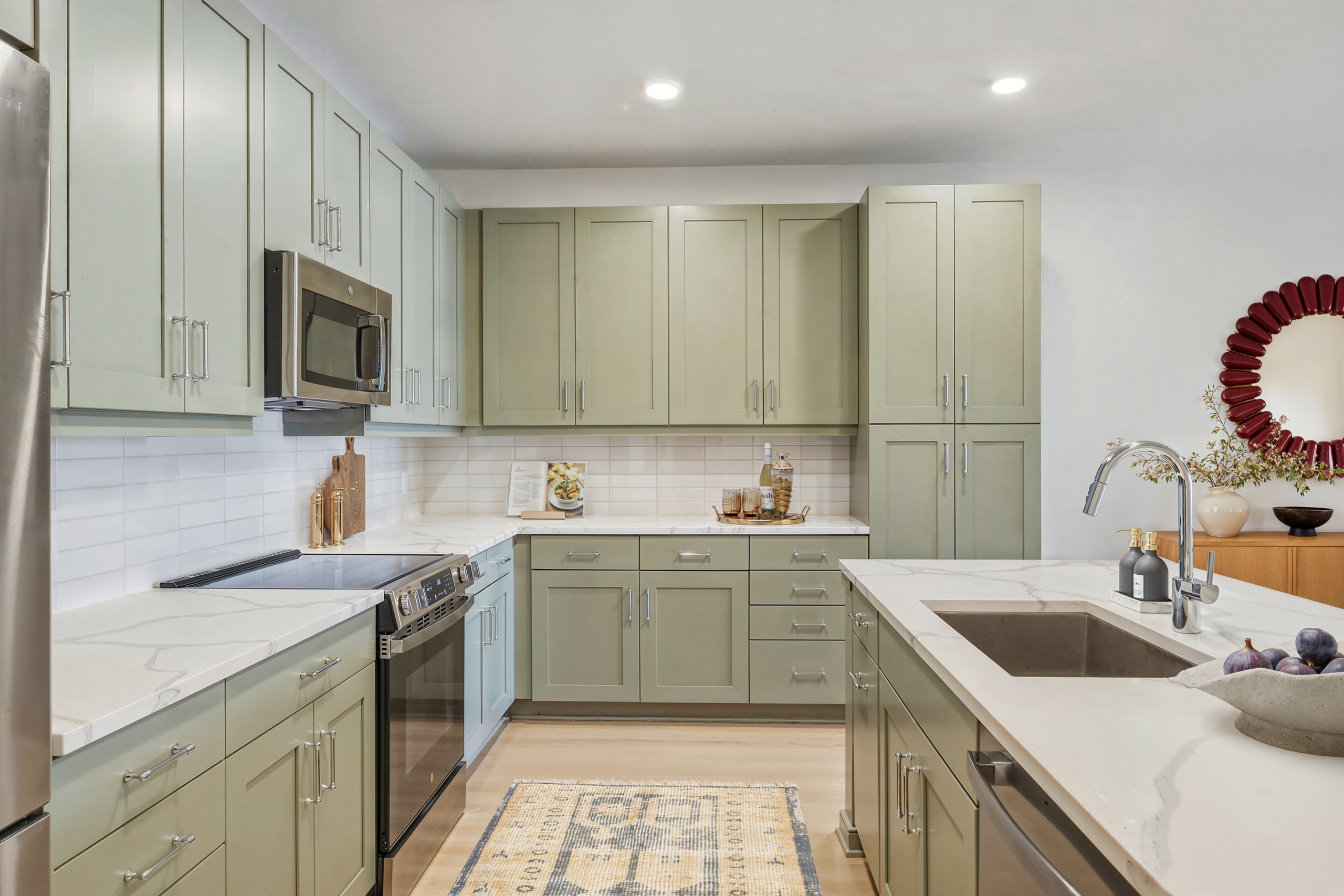 Modern kitchen with sage green shaker cabinets, white quartz countertops, stainless steel appliances, subway tile backsplash, and light wood flooring.