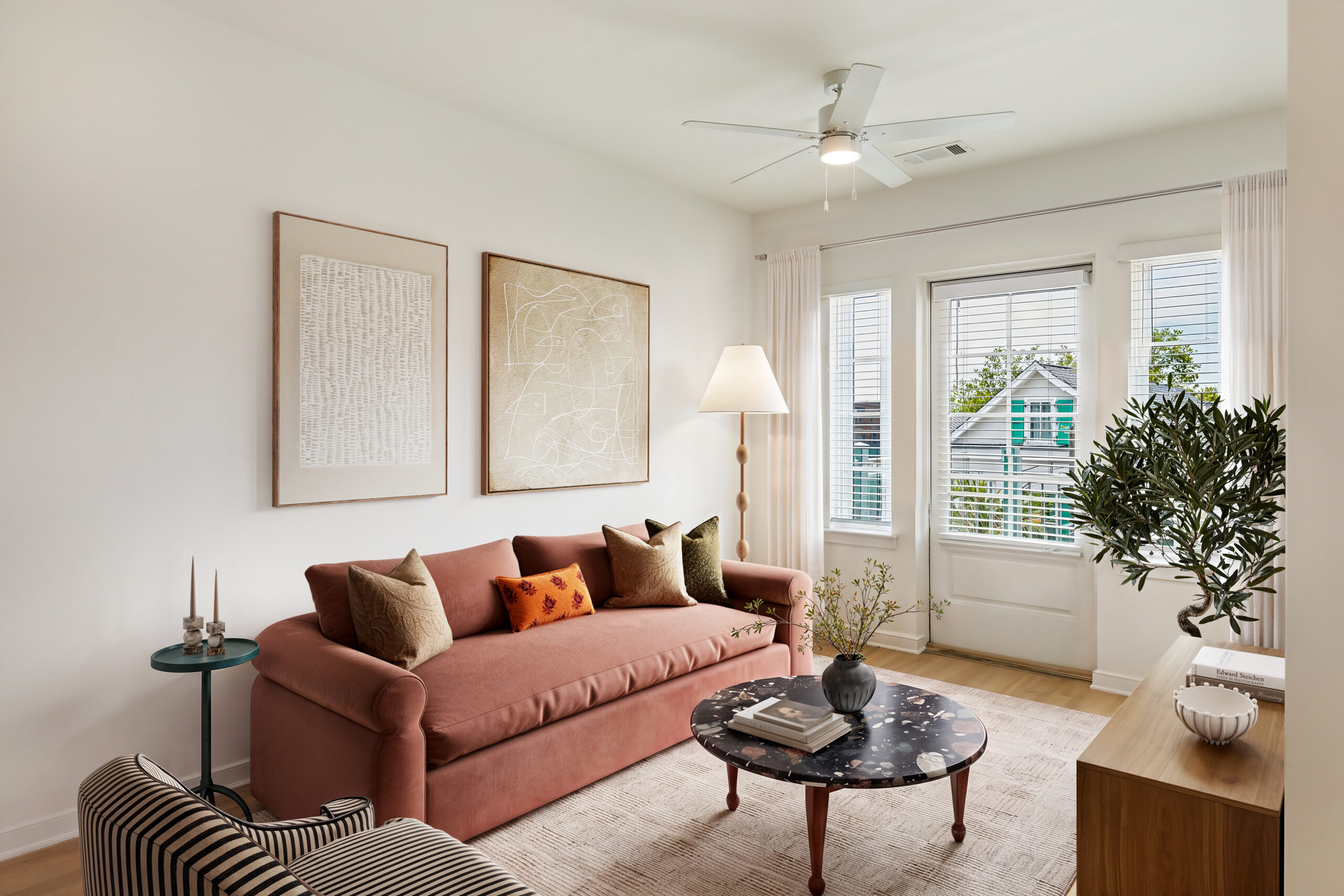 Modern living room with a blush pink sofa, round black coffee table, large windows, natural light, neutral décor, indoor plant, and contemporary minimalist design.