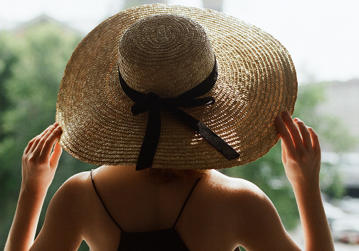 A view from behind of a woman wearing a wide-brimmed straw hat with a black ribbon tied in a bow. She is wearing a black top with thin straps and has her hands raised to the brim of the hat. The background is a soft-focus view of green trees.