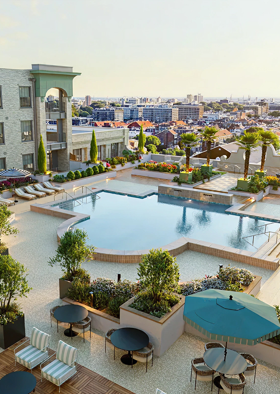 The rooftop pool area of The Laurel, featuring a uniquely shaped swimming pool surrounded by a light-colored stone deck. The space is elegantly landscaped with palm trees and flower beds, offering various seating options including striped lounge chairs and patio tables with umbrellas. In the background, an expansive view of the city skyline stretches toward the horizon under a clear sky.