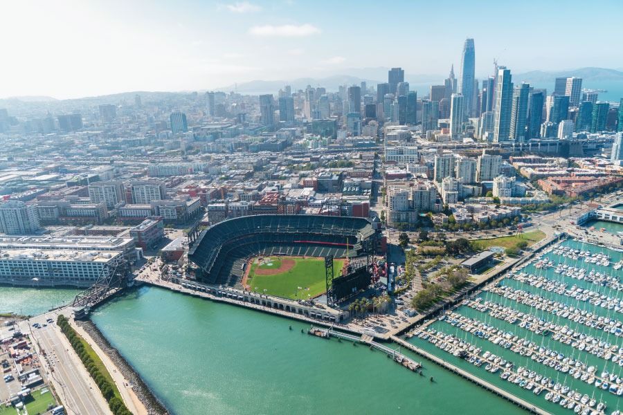 A breathtaking aerial view of a sun-drenched coastal city. In the foreground, a large, modern professional baseball stadium sits at the water's edge, characterized by its dark green seating and perfectly manicured grass field. To the right, a densely packed marina is filled with hundreds of white sailboats and yachts. The background transitions into a massive urban skyline with glass-walled skyscrapers and diverse architectural styles stretching toward distant rolling hills. The scene captures the intersection of professional sports, luxury maritime culture, and high-density metropolitan living under a clear, bright sky.
