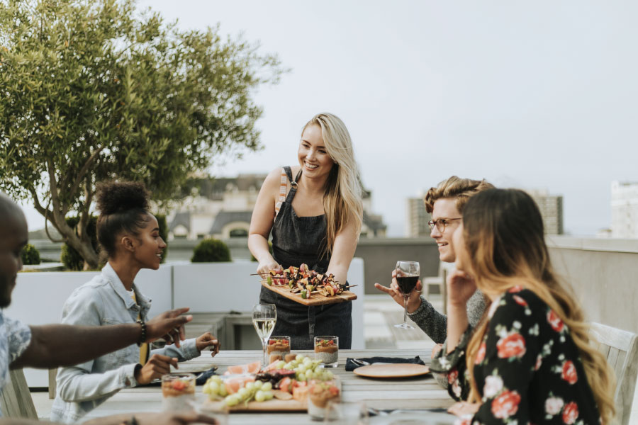 A bright, candid outdoor shot of a diverse group of friends enjoying a meal on a sun-drenched rooftop terrace. In the center, a smiling woman in a dark denim apron serves a wooden platter of grilled skewers to guests seated around a rustic wooden table. The table is spread with a vibrant feast including fresh fruit, grapes, and glasses of white and red wine. The atmosphere is relaxed and joyful, set against a backdrop of soft greenery and a blurred metropolitan skyline, embodying a lifestyle of connection, leisure, and refined community living.