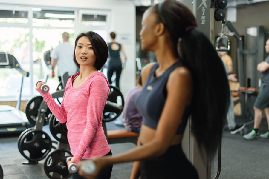 A candid, medium shot of two women working out in a bright, modern fitness center. In the foreground, a woman with short dark hair and a pink athletic top is performing bicep curls with dumbbells, looking toward her companion. Beside her, another woman with a long dark ponytail is partially visible in profile, wearing a navy blue sports top. The background is softly blurred, showing professional-grade gym equipment, including treadmills and weight racks, along with other gym-goers near large windows that let in plenty of natural light. The image captures a sense of community, health, and a high-energy lifestyle.