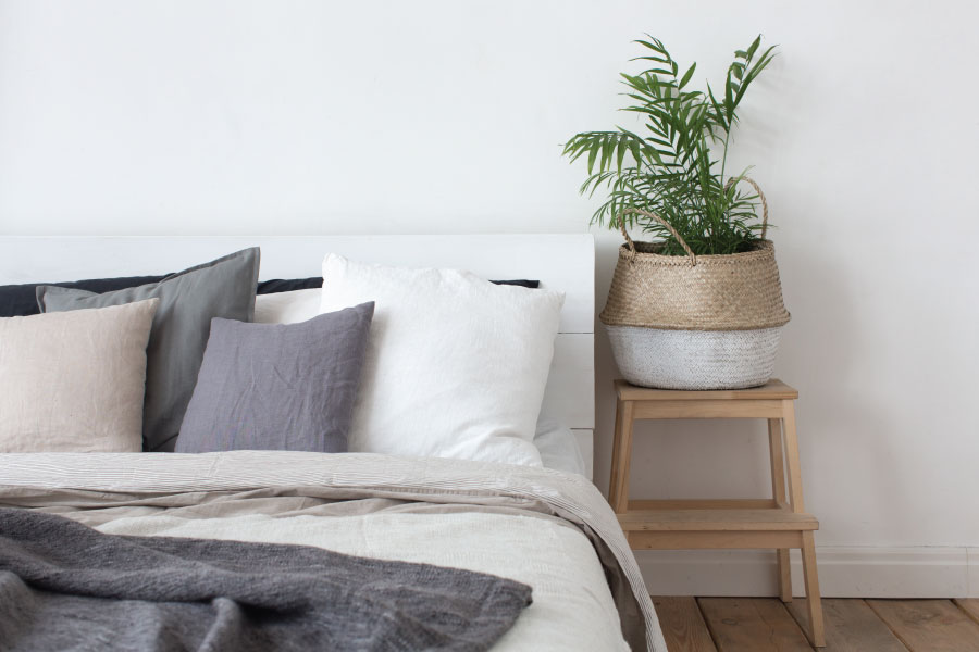 A serene and minimalist bedroom detail featuring a bed with layered linens in a neutral palette of white, cream, and charcoal grey. Several linen accent pillows in muted tones of beige and slate are propped against a simple white headboard. To the right, a small light-wood step stool serves as a nightstand, holding a woven seagrass basket planter with a vibrant green parlor palm. The scene is set against a clean white wall and light wood flooring, illuminated by soft, natural light to create a peaceful and high-end residential atmosphere.