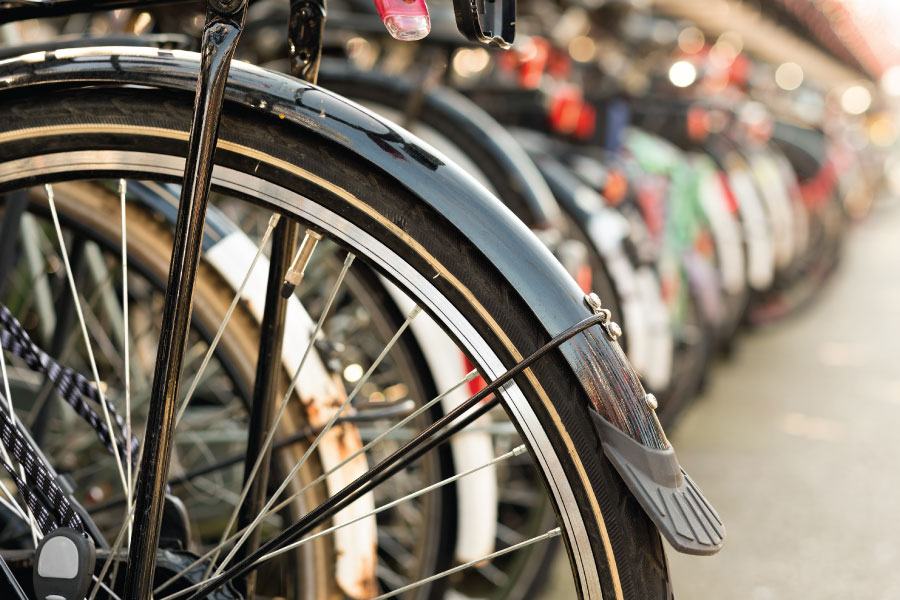 A crisp, close-up photograph of a row of parked bicycles with a shallow depth of field. The focus is sharp on the metallic black fender and spokes of the lead bike, showcasing the mechanical detail and tire tread. The rest of the bicycles trail off into a soft, blurred background, creating a rhythmic and modern urban feel. The shot emphasizes a lifestyle of mobility, active transit, and the energy of a bustling, high-density professional district.