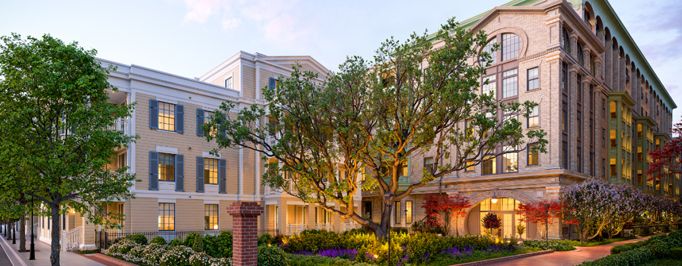 A sprawling twilight view of The Laurel, showing its multi-building residential campus. To the left, the architecture features classic cream-colored siding and blue-grey shutters, while the right side showcases the grand stone facade with a massive signature arched entryway. In the center, a large, mature tree is beautifully uplighted, anchored in a lush landscape of flowers and red maples, all connected by clean brick walkways that define the upscale atmosphere of The Laurel.