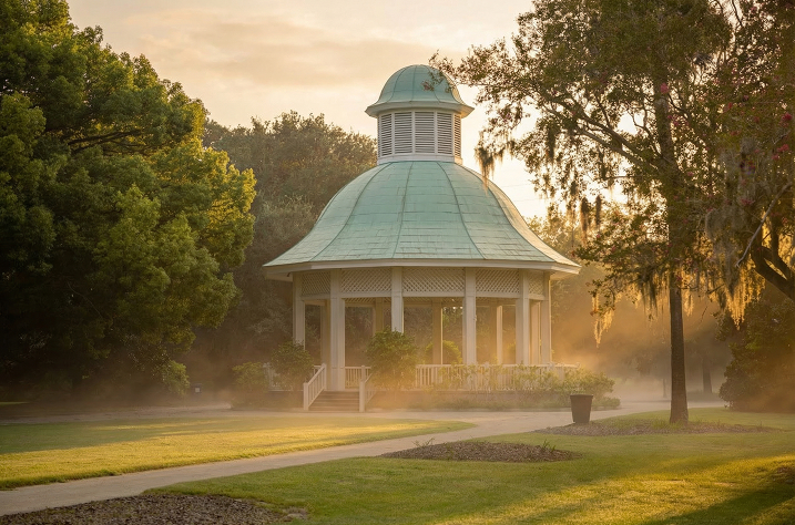 A white gazebo with a green domed roof stands in a park at sunrise. Golden sunlight and morning mist filter through the surrounding lush green trees and Spanish moss, casting a warm glow over the grass and winding paved walkway.