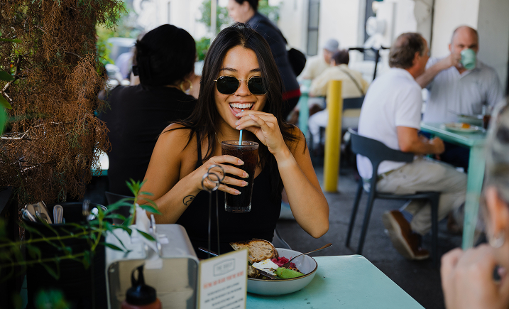A young woman with dark hair and sunglasses sits at a light teal outdoor table, smiling as she sips a dark drink through a straw. In front of her is a bowl containing avocado, toast, and eggs. The background shows a blurred outdoor patio scene with other patrons dining at nearby tables under bright, natural daylight.
