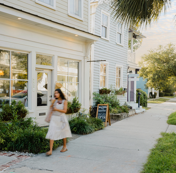 A woman walking along a sunny concrete sidewalk past a row of charming two-story wooden buildings with light-colored siding. The storefront in the foreground features large windows and a white door with a window box planter, while a chalkboard sign for 