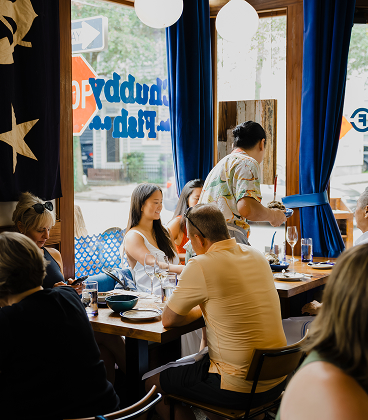 The interior of a lively, sunlit restaurant with blue curtains and wooden window frames. Patrons are seated at wooden tables, including a woman in white and a man in a yellow shirt, while a server in a floral shirt carries a plate through the dining room. Through the window, the blue 