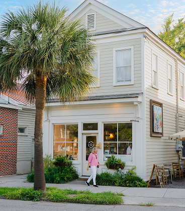 A two-story, cream-colored wooden building with a storefront on the ground floor. A woman in a pink shirt and white pants walks along the sidewalk in front of the shop, which features large glass windows and window box planters. A tall palm tree stands to the left of the building under a clear blue sky, while an outdoor seating area with a wooden A-frame sign is visible to the right.