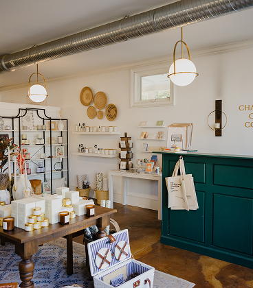 The interior of a bright boutique shop featuring a dark green service counter with canvas tote bags hanging from a hook. In the foreground, a wooden table displays various candles in white boxes and jars, while the background walls are lined with white floating shelves and black metal racks holding various retail goods. The space is illuminated by modern globe pendant lights with brass hardware and features decorative woven baskets on the wall.