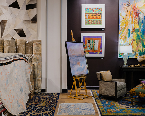 An eclectic interior design studio featuring an easel with an abstract landscape painting. The space is filled with colorful patterned rugs, a grey armchair, and a dark purple accent wall displaying framed art, including a portrait and a vibrant abstract canvas.