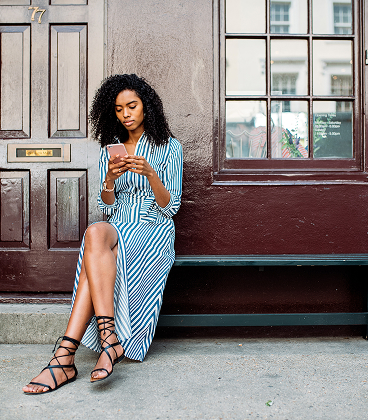 A woman with curly hair wearing a blue and white striped wrap dress and black lace-up sandals sits on a dark bench outside a building. She is looking down at a smartphone in her hands. Behind her is a dark brown textured wall and a matching wooden door with a brass mail slot and the number 