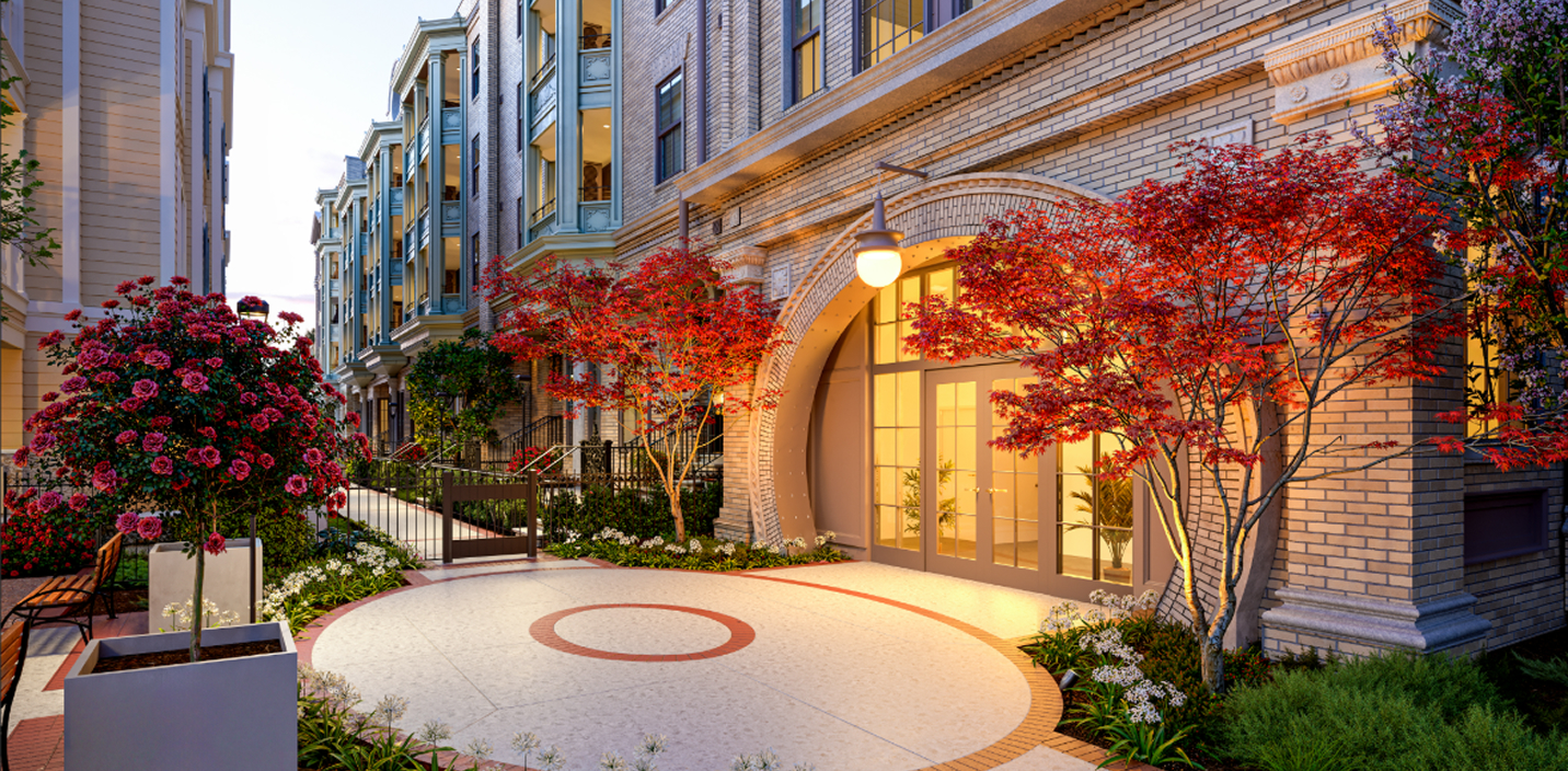 The brick exterior and arched entryway of The Laurel, featuring a circular stone patio and bright red maple trees. A glowing orb light hangs above the glass double doors, which lead into a brightly lit lobby, while a landscaped walkway with rose bushes and benches extends along the side of the building.