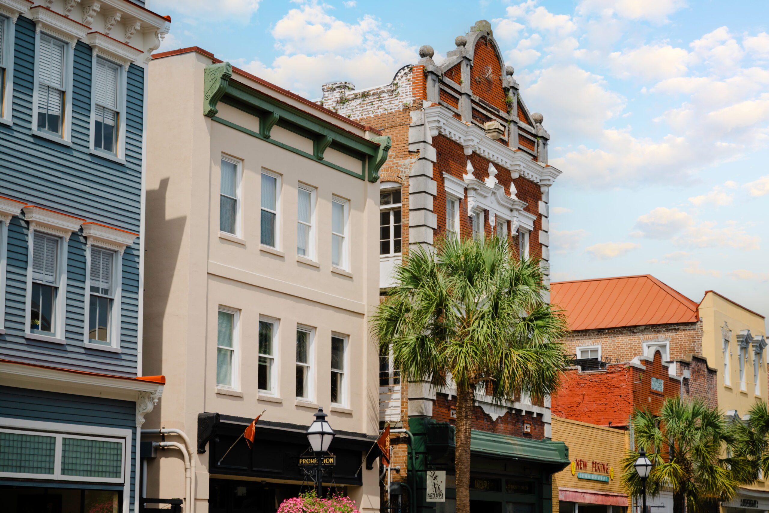A detailed street view capturing a row of four distinct, interconnected historic building facades. On the far left is a multi-story building with blue-grey horizontal clapboard siding and intricate white ornamental window pediments. To its right stands a tan, two-story building featuring prominent decorative green bracket eaves, located above black-framed ground-level storefronts with gas-style street lamps. The central and tallest building is constructed from weathered red brick, showcasing a complex white gabled pediment, ornate roofline cornices, and decorative stone quoins along its edges. A tall, textured-trunk palm tree with vibrant green fronds stands directly in front of this brick building. Further to the right, adjacent buildings feature exposed brick and pale yellow facades with varied rooflines under a bright, partly cloudy blue sky.