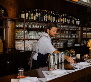 A bearded bartender in a white shirt and dark apron works behind a classic, dark wood bar. He is reaching for a small glass on the counter near several metal cocktail shakers and neatly folded white napkins. In the background, tiered shelves are densely packed with a variety of liquor bottles and rows of clear glassware.