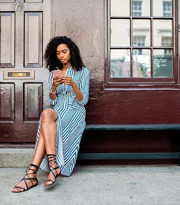 A woman with curly hair wearing a blue and white striped wrap dress and black lace-up sandals sits on a dark bench outside a building. She is looking down at a smartphone in her hands. Behind her is a dark brown textured wall and a matching wooden door with a brass mail slot and the number 