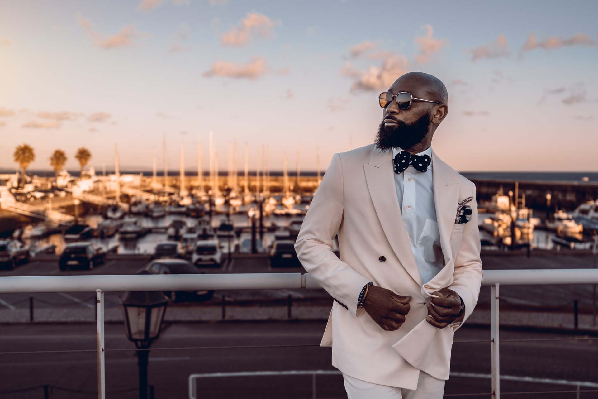 Person in suit with bowtie and sunglasses leaning against railing in front of marina.