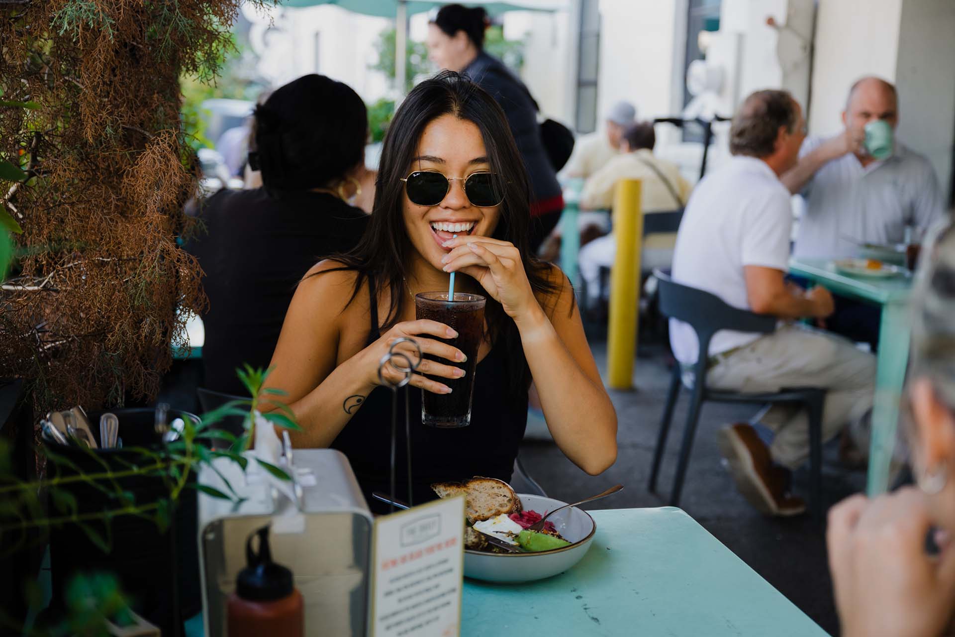 A young woman with dark hair and sunglasses sits at a light teal outdoor table, smiling as she sips a dark drink through a straw. In front of her is a bowl containing avocado, toast, and eggs. The background shows a blurred outdoor patio scene with other patrons dining at nearby tables under bright, natural daylight.