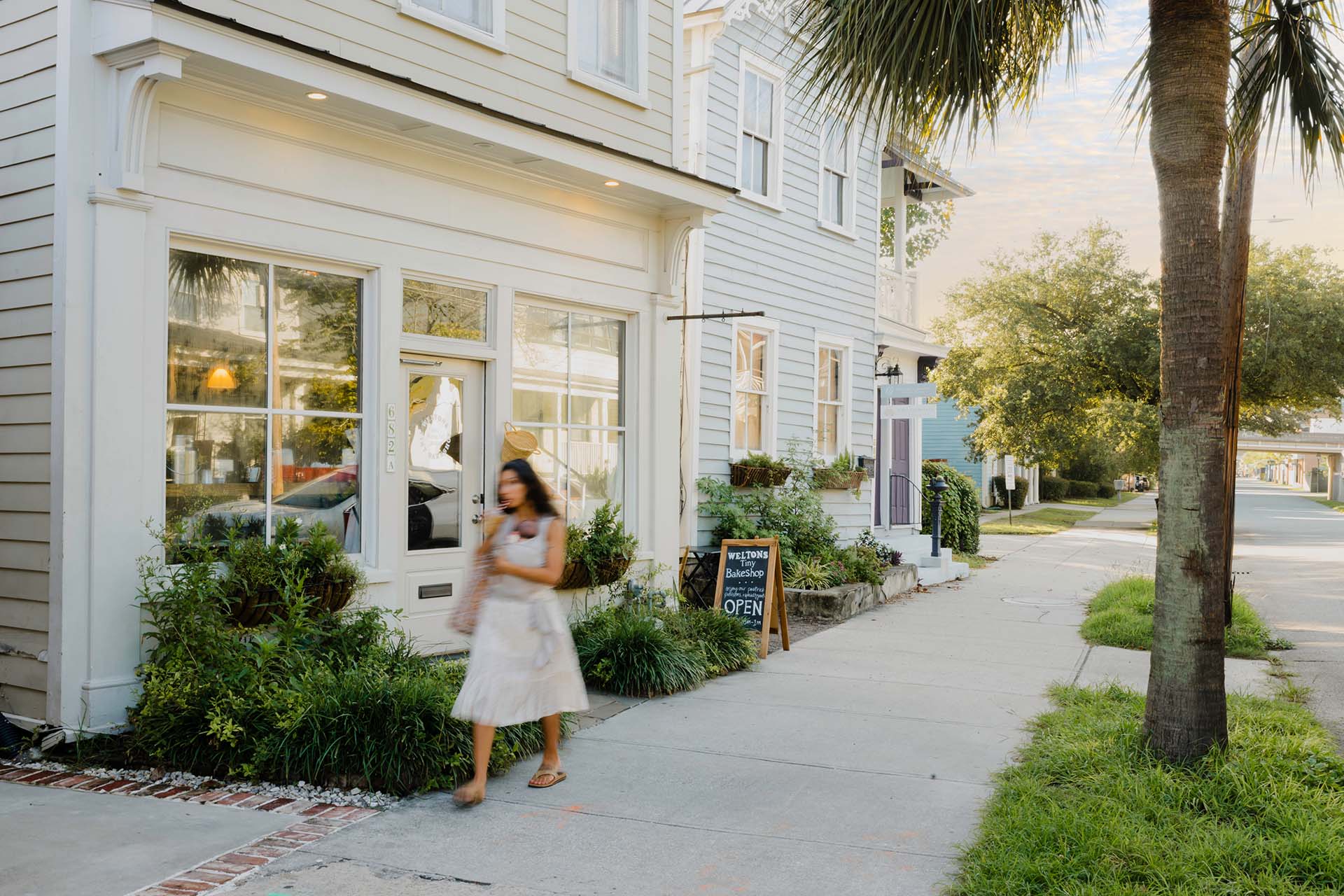 A woman walking along a sunny concrete sidewalk past a row of charming two-story wooden buildings with light-colored siding. The storefront in the foreground features large windows and a white door with a window box planter, while a chalkboard sign for 