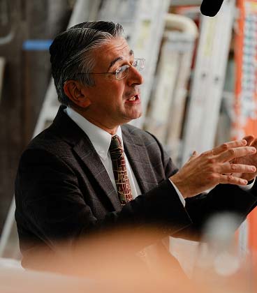 A middle-aged man with grey hair and glasses wearing a dark patterned suit, a white shirt, and a decorative tie. He is captured in profile, speaking and gesturing with both hands. The background is softly blurred, showing industrial-style elements like ladders and shelving.