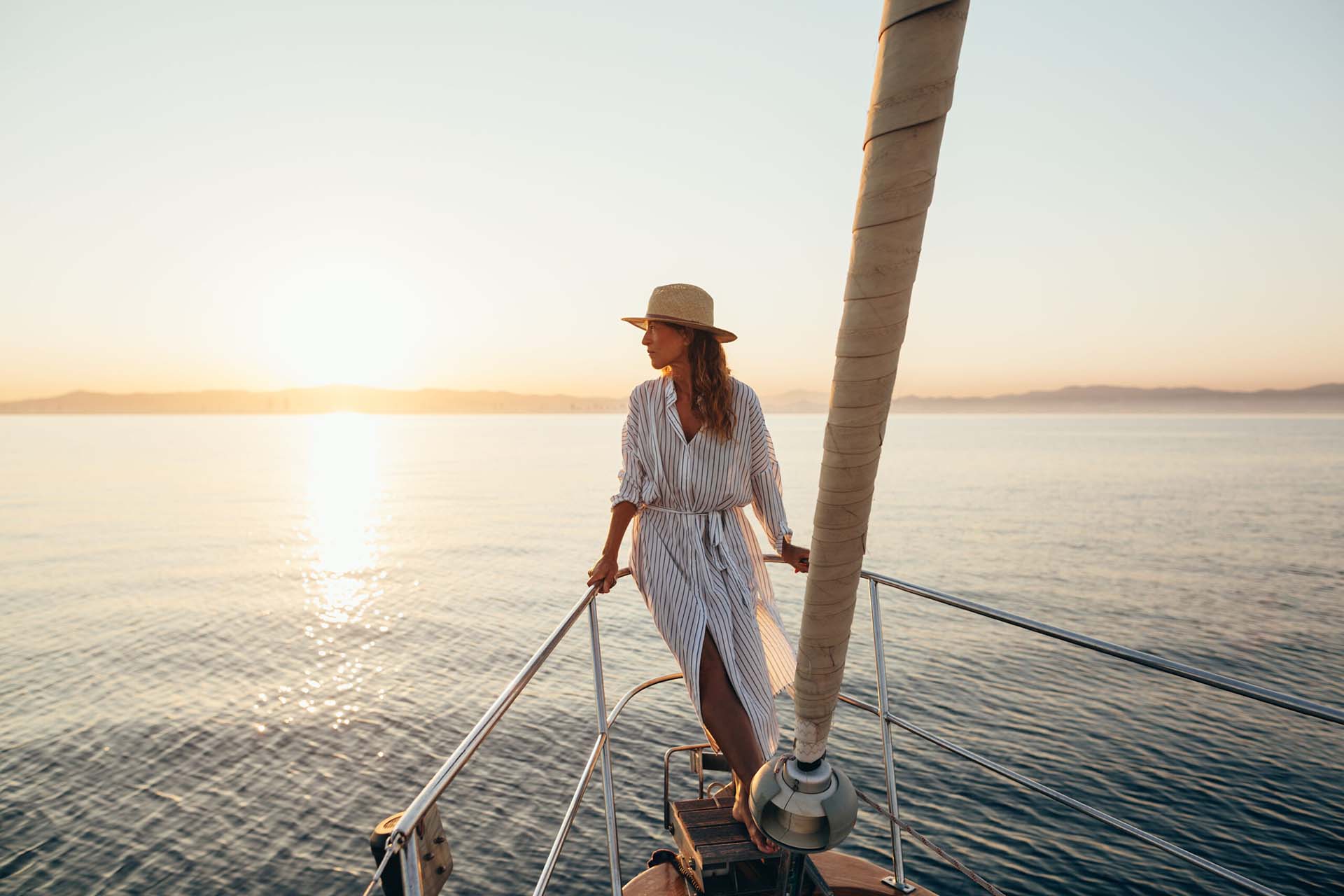 A wide, cinematic shot taken from the bow of a sailboat at sunrise or sunset. A woman stands gracefully at the front of the boat, leaning against the metal railing and looking out toward the distant, hazy horizon. She is wearing a wide-brimmed straw sun hat and a long, breezy white-and-grey striped linen dress that caught in the light wind. The calm, glistening water reflects the golden glow of the sun, which sits low in a clear, pale sky. The foreground features the base of the sailboat’s mast and taut rigging, while a soft coastline is visible in the far distance, creating an aspirational atmosphere of peace, success, and leisure.