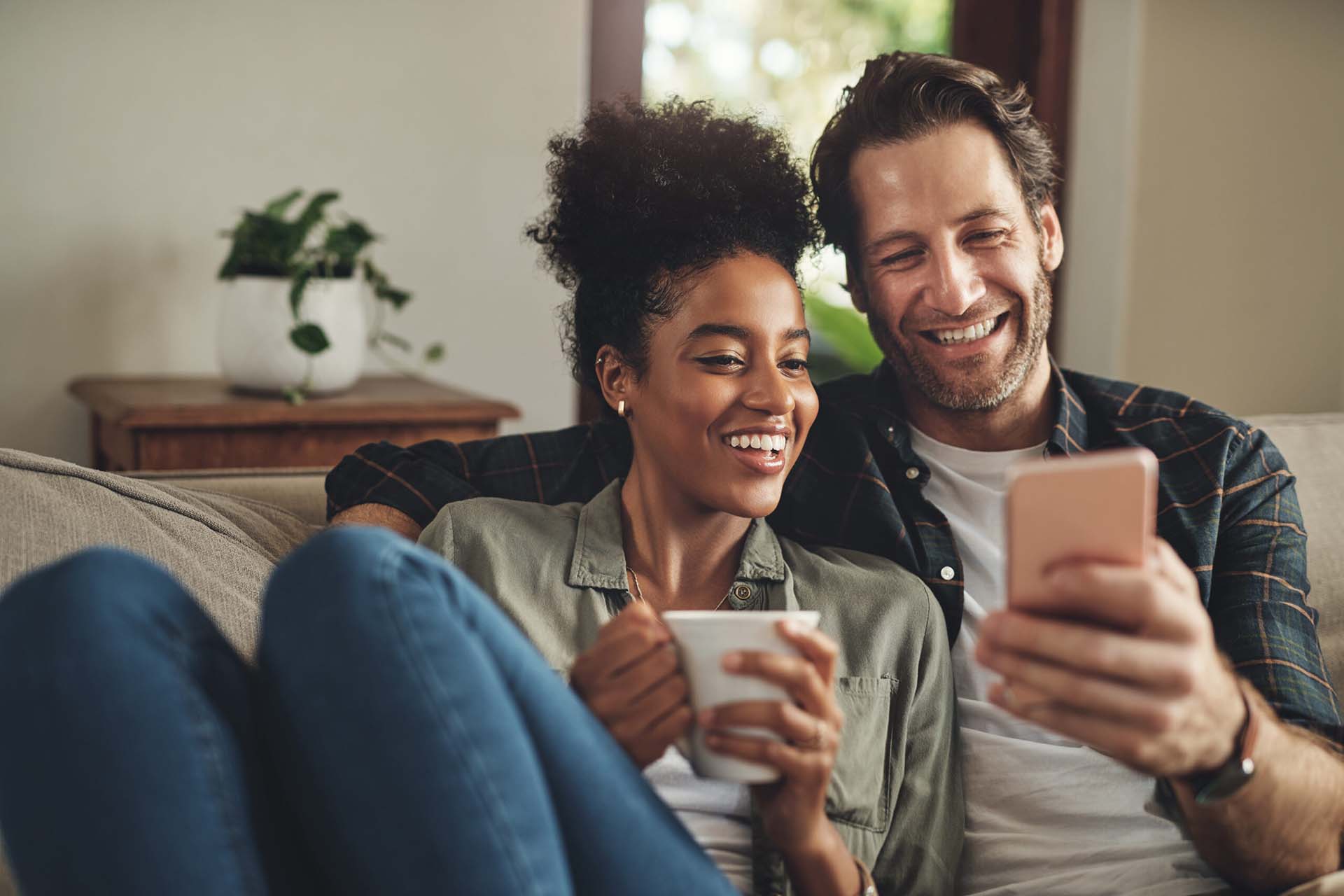 Smiling couple sitting on a couch together with coffee and a smart phone.